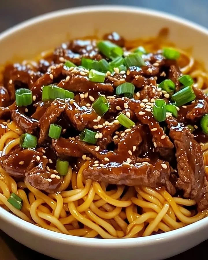Plate of sticky beef noodles topped with sesame seeds and herbs.